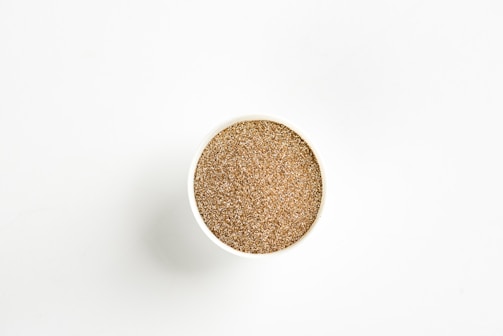 A round container filled with a granular, sand-like substance is centered against a plain white background. The contents appear to have a uniform brown color with a coarse texture.