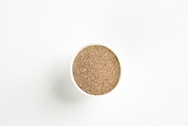 A round container filled with a granular, sand-like substance is centered against a plain white background. The contents appear to have a uniform brown color with a coarse texture.
