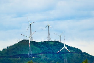 Several wind turbines stand atop a green hill, surrounded by a clear sky filled with clouds. The turbines are evenly spaced and exhibit modern engineering with metal support structures, contributing to a clean energy landscape.