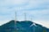 Several wind turbines stand atop a green hill, surrounded by a clear sky filled with clouds. The turbines are evenly spaced and exhibit modern engineering with metal support structures, contributing to a clean energy landscape.