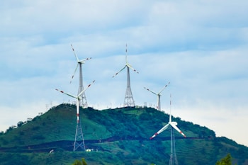 Several wind turbines stand atop a green hill, surrounded by a clear sky filled with clouds. The turbines are evenly spaced and exhibit modern engineering with metal support structures, contributing to a clean energy landscape.
