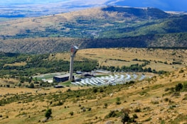 A large solar power installation is situated in a rolling landscape with a tall tower at the center, surrounded by numerous solar panels arranged in a circular pattern. The surrounding rural terrain features grassy hills and sparsely distributed trees, with distant mountains visible under a clear blue sky.