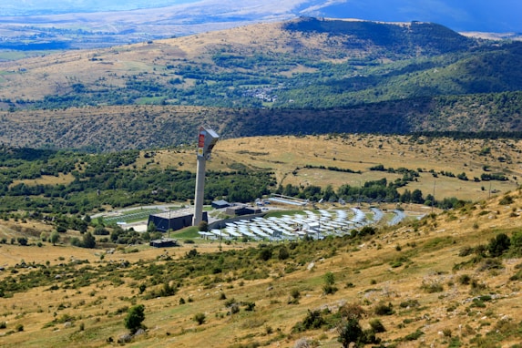 A large solar power installation is situated in a rolling landscape with a tall tower at the center, surrounded by numerous solar panels arranged in a circular pattern. The surrounding rural terrain features grassy hills and sparsely distributed trees, with distant mountains visible under a clear blue sky.