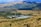 A large solar power installation is situated in a rolling landscape with a tall tower at the center, surrounded by numerous solar panels arranged in a circular pattern. The surrounding rural terrain features grassy hills and sparsely distributed trees, with distant mountains visible under a clear blue sky.
