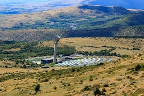 A large solar power installation is situated in a rolling landscape with a tall tower at the center, surrounded by numerous solar panels arranged in a circular pattern. The surrounding rural terrain features grassy hills and sparsely distributed trees, with distant mountains visible under a clear blue sky.