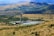 A large solar power installation is situated in a rolling landscape with a tall tower at the center, surrounded by numerous solar panels arranged in a circular pattern. The surrounding rural terrain features grassy hills and sparsely distributed trees, with distant mountains visible under a clear blue sky.