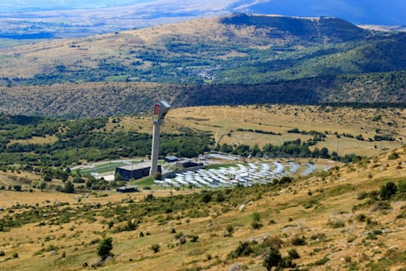A large solar power installation is situated in a rolling landscape with a tall tower at the center, surrounded by numerous solar panels arranged in a circular pattern. The surrounding rural terrain features grassy hills and sparsely distributed trees, with distant mountains visible under a clear blue sky.