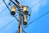 A wooden utility pole supports three cylindrical transformers, surrounded by numerous black electrical wires against a clear blue sky.