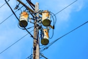 A wooden utility pole supports three cylindrical transformers, surrounded by numerous black electrical wires against a clear blue sky.