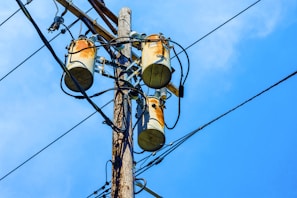 A wooden utility pole supports three cylindrical transformers, surrounded by numerous black electrical wires against a clear blue sky.
