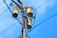 A wooden utility pole supports three cylindrical transformers, surrounded by numerous black electrical wires against a clear blue sky.
