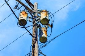 A wooden utility pole supports three cylindrical transformers, surrounded by numerous black electrical wires against a clear blue sky.