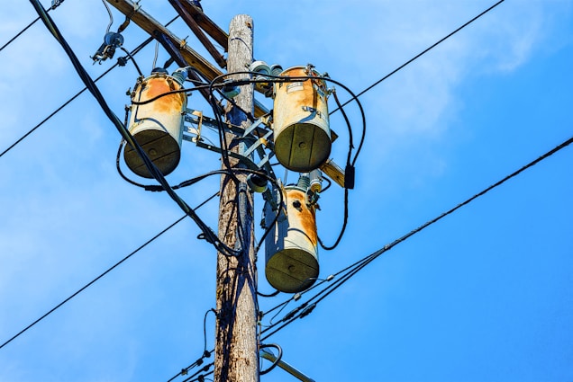 A wooden utility pole supports three cylindrical transformers, surrounded by numerous black electrical wires against a clear blue sky.