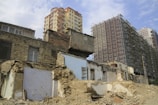 A partially demolished building with exposed interiors and crumbling walls is situated in front of taller, modern structures under construction. The contrast between old, damaged architecture and new, developing high-rises is prominent.