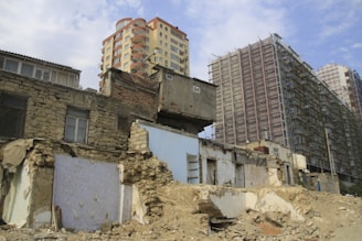 A partially demolished building with exposed interiors and crumbling walls is situated in front of taller, modern structures under construction. The contrast between old, damaged architecture and new, developing high-rises is prominent.