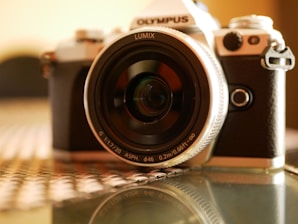 A close-up shot of a camera with a large lens, displaying the brand names 'Olympus' and 'Lumix.' The camera is placed on a glass surface, and the background is softly blurred, creating a shallow depth of field effect.