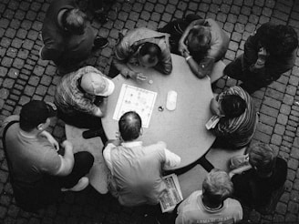 A group of people are gathered around a circular table, playing a board game. The scene is viewed from above and is in black and white, adding a timeless feel. The game board is positioned in the center, with various pieces and players appearing engaged. The surrounding cobblestone ground suggests an outdoor setting.