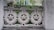 Ornate concrete design features circular patterns with flower-like motifs against a backdrop of green foliage and yellow flowers. The architectural element appears weathered, contributing to a rustic aesthetic.