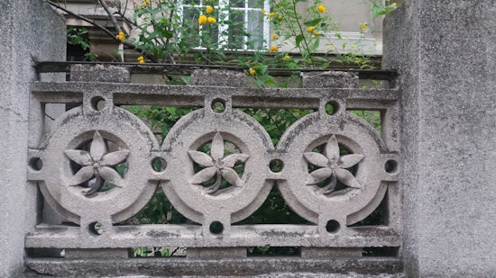 Ornate concrete design features circular patterns with flower-like motifs against a backdrop of green foliage and yellow flowers. The architectural element appears weathered, contributing to a rustic aesthetic.