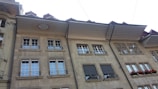 A row of classic European-style buildings with multiple windows and ornate wrought iron balcony railings. The facade features a stone material with decorative elements, including a date inscribed on one of the upper sections. Bright red flowers adorn one of the windows.