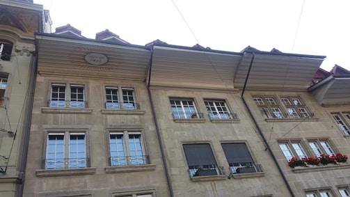 A row of classic European-style buildings with multiple windows and ornate wrought iron balcony railings. The facade features a stone material with decorative elements, including a date inscribed on one of the upper sections. Bright red flowers adorn one of the windows.