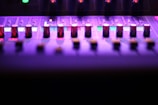 A close-up view of a sound mixing console, featuring multiple knobs and sliders under purple and pink lighting. The image captures the sleek and modern design, highlighting the metal sheen of the knobs. Small illuminated sections add a futuristic feel to the overall atmosphere.