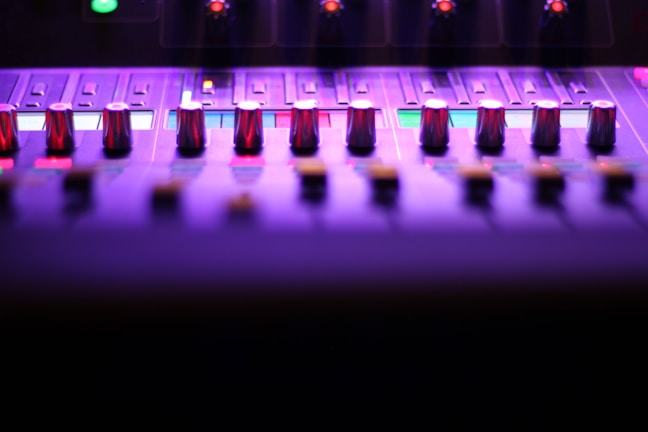 A close-up view of a sound mixing console, featuring multiple knobs and sliders under purple and pink lighting. The image captures the sleek and modern design, highlighting the metal sheen of the knobs. Small illuminated sections add a futuristic feel to the overall atmosphere.