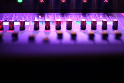 A close-up view of a sound mixing console, featuring multiple knobs and sliders under purple and pink lighting. The image captures the sleek and modern design, highlighting the metal sheen of the knobs. Small illuminated sections add a futuristic feel to the overall atmosphere.