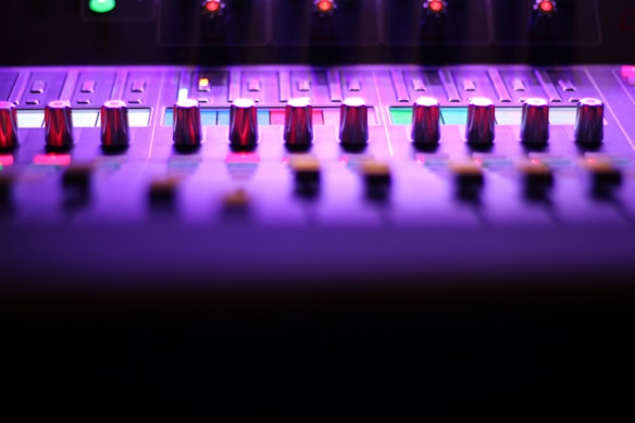 A close-up view of a sound mixing console, featuring multiple knobs and sliders under purple and pink lighting. The image captures the sleek and modern design, highlighting the metal sheen of the knobs. Small illuminated sections add a futuristic feel to the overall atmosphere.