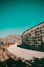 A curved wall of niches, possibly a mausoleum or columbarium, curves alongside a dirt path. This structure is surrounded by a mountainous landscape under a vibrant teal sky. On the left, the path is bordered by dried foliage and small trees. The setting feels remote and tranquil.
