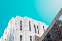 A clean and clear architectural composition featuring an impressive art deco building with white stone facades and tall, narrow windows. Adjacent to it is a modern building with a glass facade, reflecting light and adding a contemporary element. The background is a bright, vibrant blue sky, enhancing the contrast between the buildings.