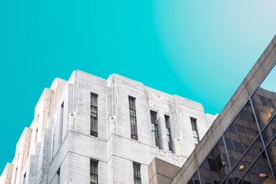 A clean and clear architectural composition featuring an impressive art deco building with white stone facades and tall, narrow windows. Adjacent to it is a modern building with a glass facade, reflecting light and adding a contemporary element. The background is a bright, vibrant blue sky, enhancing the contrast between the buildings.