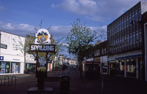 A street scene featuring a sign with the word 'SPALDING' and an emblem above it. Trees are lining the street, which is flanked by buildings. There are shops in the background, including a pharmacy. A phone booth and bicycle racks are also visible, creating an urban landscape under a partly cloudy sky.