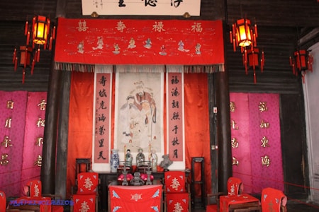 A traditional Chinese shrine with vibrant red and pink decorative elements includes lanterns, banners with Chinese calligraphy, and an ornate tapestry depicting traditional figures. Various objects, like vases and incense holders, are arranged on a wooden altar.