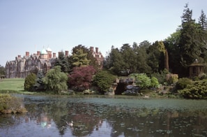 A grand, historic mansion stands on a serene estate surrounded by lush greenery and trees. A peaceful pond with waterfowl in the foreground reflects the blue sky and the mansion. Red and green foliage adds color to the landscape.