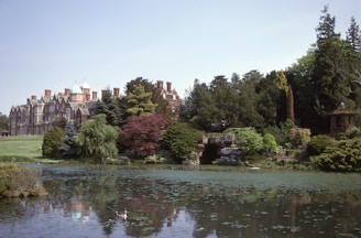 A grand, historic mansion stands on a serene estate surrounded by lush greenery and trees. A peaceful pond with waterfowl in the foreground reflects the blue sky and the mansion. Red and green foliage adds color to the landscape.