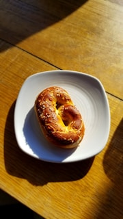 A sesame seed-covered bagel is placed on a white plate. The plate is positioned on a wooden table, and sunlight casts shadows across the surface.
