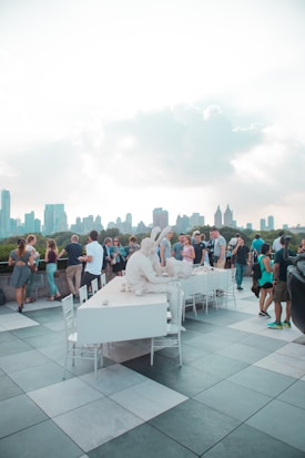 A group of people gathered on a rooftop with a scenic city skyline in the background. Centered in the foreground is a white sculptural installation of two seated figures at a long table. The scene is lively, suggesting a social event or gathering in an open-air setting with a mosaic-tiled floor.