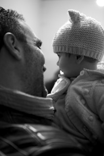 A black and white photograph captures a tender moment between an adult and a baby. The adult, seen from the side, has a warm expression while holding the baby. The baby is wearing a knitted hat with small ears and is dressed in a cozy sweater. The two are engaged in an intimate gaze, conveying a sense of affection and connection.
