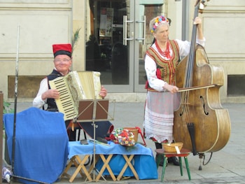 Two musicians in traditional clothing are performing on a street. One is playing an accordion while the other is playing a double bass. Both are dressed in vibrant and decorative attire with traditional patterns and accessories. They are surrounded by baskets and a small table covered with a blue cloth.