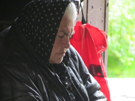 An elderly person is sitting quietly with eyes closed, wearing a dark polka-dot headscarf and a black jacket. A bright red bag hangs on the door in the background, contrasting with the muted tones. The scene suggests a sense of solitude.