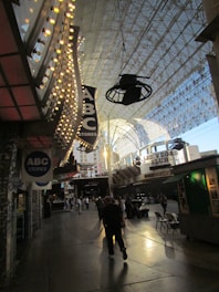 A bustling street scene under a vast canopy with decorative lights. People walk along the sidewalk below signs for ABC Stores and Las Vegas Club. The area appears lively with a blend of modern and retro aesthetics.
