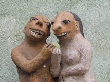 Two clay sculptures with exaggerated facial features are depicted, closely positioned against a textured green background. The figures are crafted with earthy tones and have grinning expressions, with one holding a small green object between them.