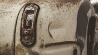 A close-up view of an old, rusted car with significant wear to its paint. The tail light casing is broken, and the surface is covered with rust and dirt, indicating long-term exposure to the elements.