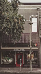 A storefront with the sign 'MAGNA CARTA' above the entrance. There is a red door at the center with a parking meter in front and a streetlamp to the side. The building has a dark, textured facade with a large tree covering part of the upper area.