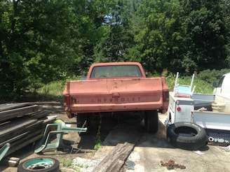 An old, rusted Chevrolet pickup truck is parked on a concrete surface surrounded by various items such as scattered tires, a stack of wooden planks, and plastic chairs. The area is partly overgrown with greenery, with tall trees in the background.