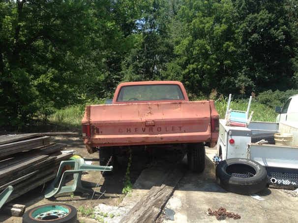 An old, rusted Chevrolet pickup truck is parked on a concrete surface surrounded by various items such as scattered tires, a stack of wooden planks, and plastic chairs. The area is partly overgrown with greenery, with tall trees in the background.