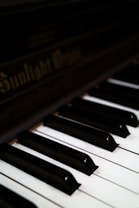Close-up view of a piano keyboard showing several black and white keys. The focus is on the keys with the brand name faintly visible on the darker wooden surface above them.