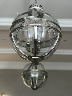 A spherical, metallic and glass ceiling light fixture with intricate detailing is hanging from the ceiling. The fixture has a reflective surface and houses several light bulbs inside. The background is a simple, neutral-toned ceiling.