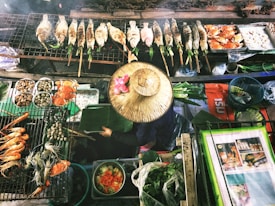 A variety of grilled seafood and other dishes are displayed on a grill, including fish, shrimp, and crabs. A person wearing a conical hat decorated with pink flowers is tending to the food, seated among vibrant ingredients like greens and vegetables. The scene suggests a market or street food setting filled with an abundance of fresh seafood and produce.
